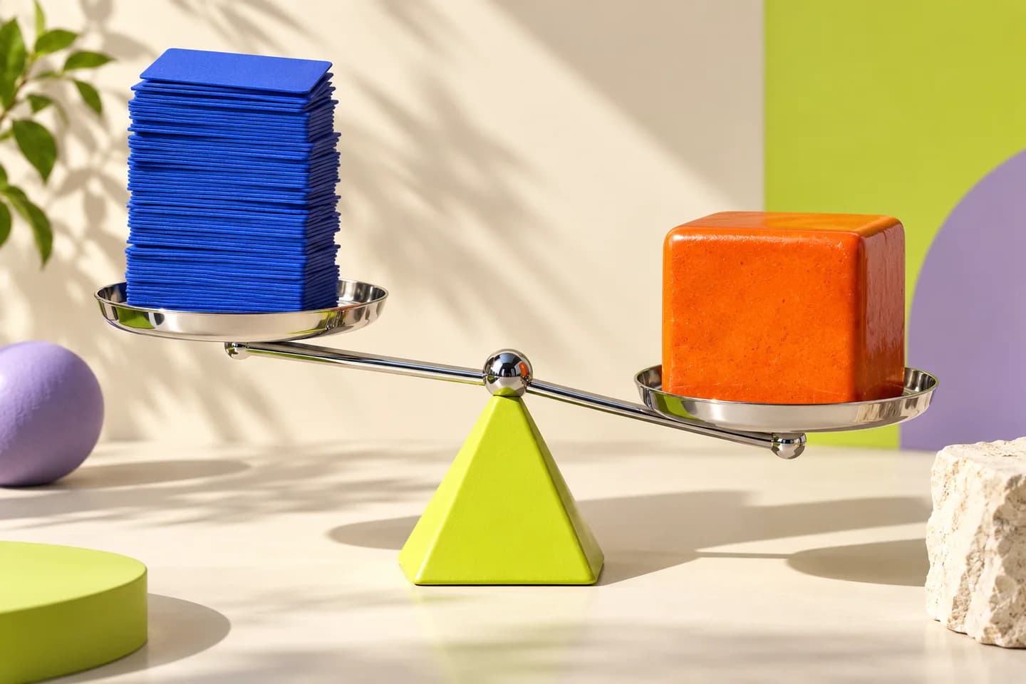 A chrome balance weighs a stack of cobalt cards against an orange block on a lime fulcrum